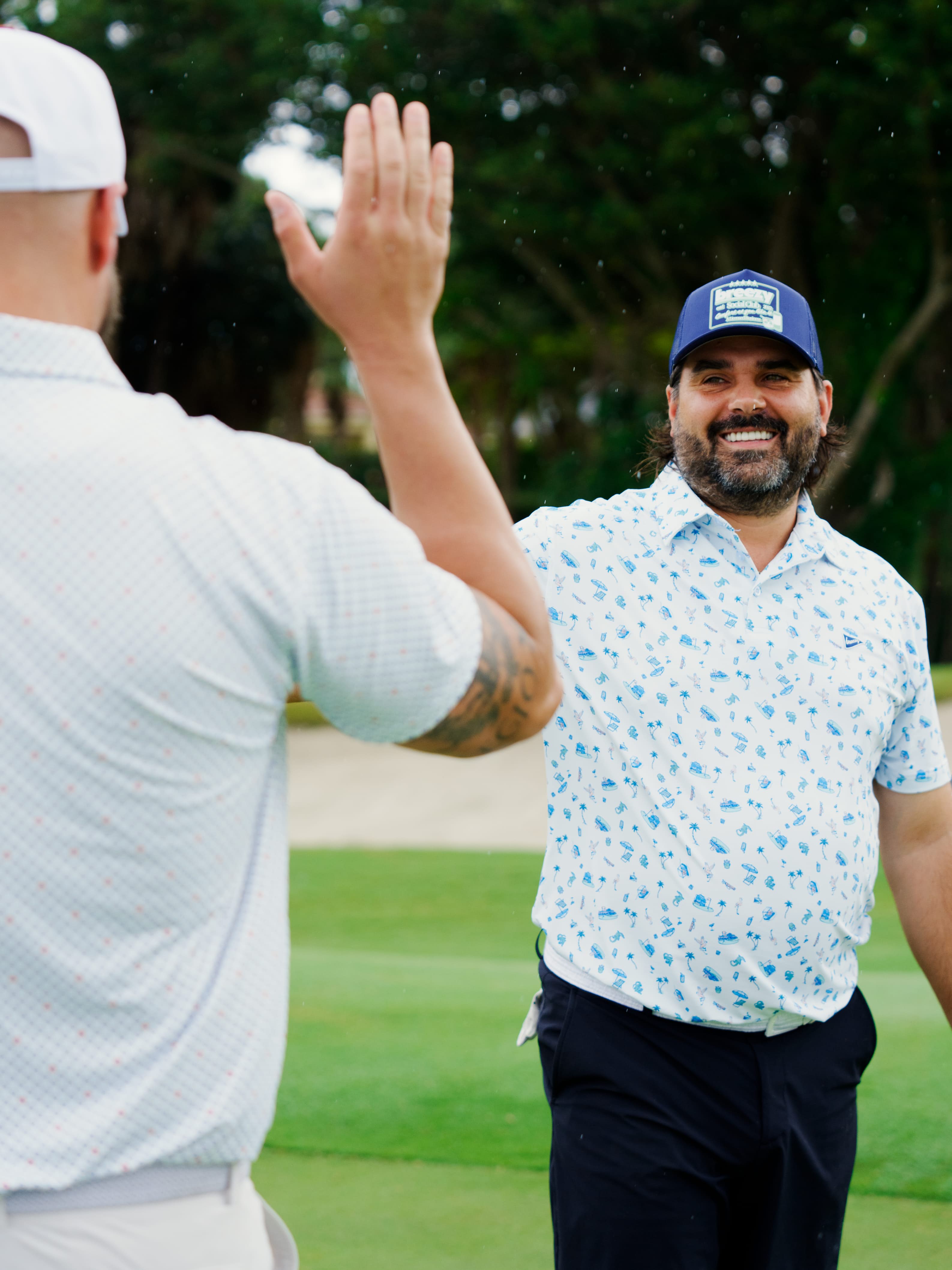 Two men on a golf course high-fiving. The man on the right is wearing a blue patterned short-sleeve polo shirt, dark pants, and a blue cap, while the man on the left is seen from behind wearing a white polo shirt and a white cap.